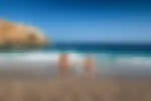 Zwei Erwachsene und ein Kind sitzen am Kiesstrand im seichten Wasser vor einer Meeresbucht mit Felsenküste