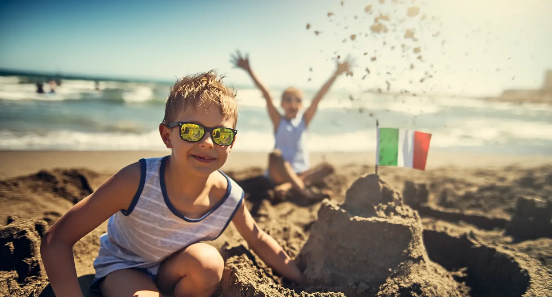 Alt-Tag: Kinder bauen am Strand in Italien eine Sandburg mit kleiner Italien-Flagge – Sommerurlaub am Meer