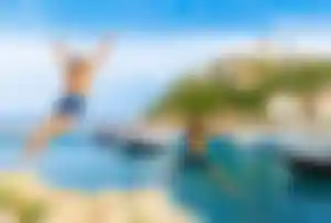 Zwei Personen springen von einer felsigen Küste in einen kleinen Hafen mit Booten und einem historischen Küstenort im Hintergrund