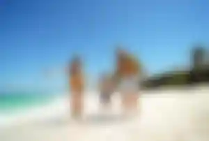 Drei Personen laufen Hand in Hand über einen hellen Sandstrand am Meer, im Hintergrund stehen Palmen und Strandhäuser.