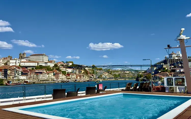 Pooldeck eines Flusskreuzfahrtschiffs mit kleinem Swimmingpool, Liegestühlen und Blick auf eine Stadt mit Brücke entlang des Flusses.