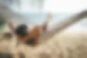 Person liegt entspannt in einer Hängematte zwischen Palmen an einem Sandstrand mit Blick auf das Meer