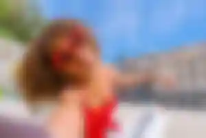 Person macht ein Selfie im Freien mit ausgestrecktem Arm, trägt auffällige Herz-Sonnenbrille und sitzt vor Gebäuden in einer Stadt bei sonnigem Wetter