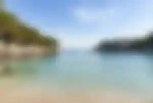 Ruhige, kleine Bucht bei Cala d’Or auf Mallorca mit hellem Sandstrand, klarem Wasser und felsiger Küste mit Pinien und Häusern im Hintergrund.