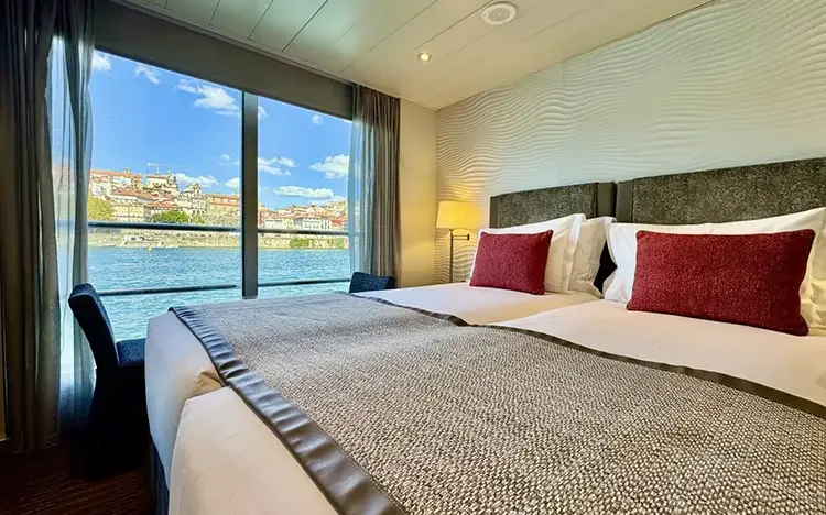 Modern eingerichtete Kabine auf einem Flusskreuzfahrtschiff mit Doppelbett und großem Panoramafenster mit Blick auf den Fluss und eine Stadt.
