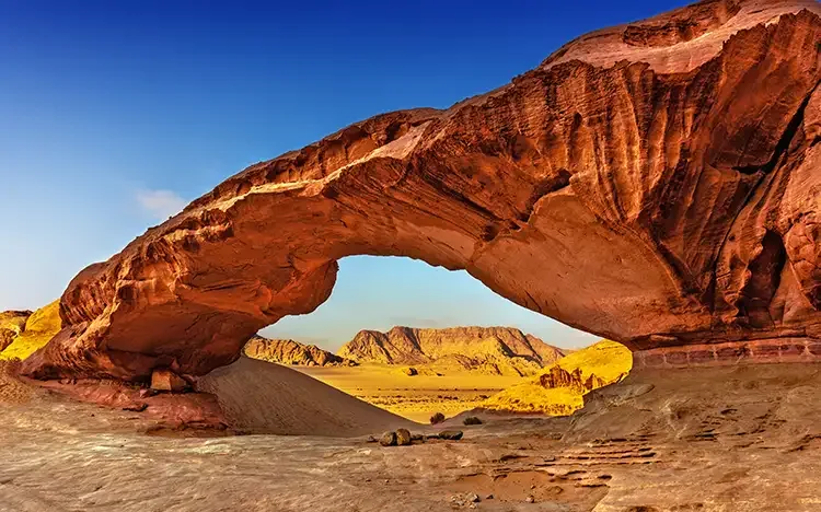 Natürliches Felsentor in der Wadi-Rum-Wüste mit rotem Sand und Bergen im Hintergrund.