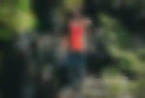 Person mit rotem Rucksack steht auf einem felsigen Weg in einer grünen, bewachsenen Landschaft und blickt in die Natur