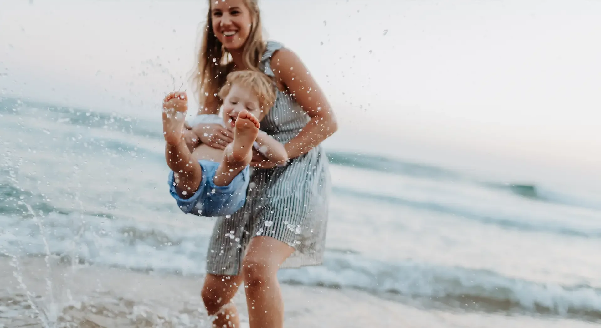 Person trägt ein Kind am Strand durch das flache Meerwasser, während Wasser spritzt und Wellen im Hintergrund zu sehen sind