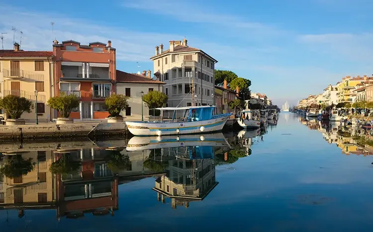 Kanal in Grado mit Fischerboot, ruhigem Wasser mit Spiegelungen und bunten Häusern unter blauem Himmel