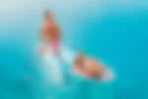 Erwachsene Person steht auf einem Stand-up-Paddle-Board in klarem, türkisem Wasser, während zwei Kinder vorne auf dem Board sitzen und mitfahren.