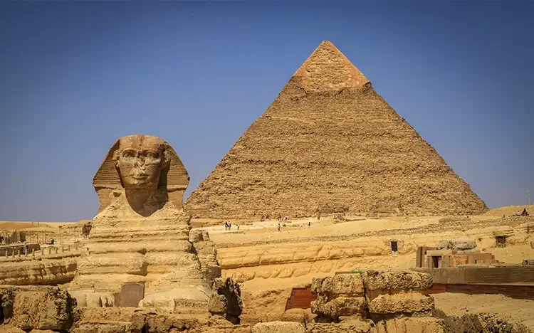 Die Große Sphinx von Gizeh vor einer der Pyramiden, umgeben von Wüstenlandschaft und antiken Steinbauten.
