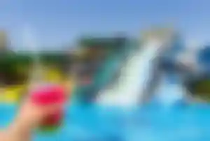 Bunter Cocktail wird vor einem Pool mit großen Wasserrutschen in einer Clubanlage in der Türkei gehalten, im Hintergrund ein moderner Aquapark.