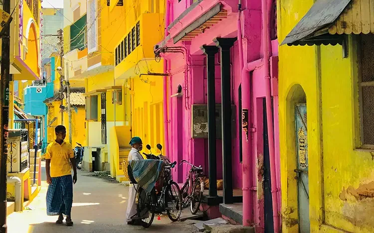 Enge Straße mit leuchtend gelben und pinken Gebäuden, in der Personen neben Fahrrädern stehen