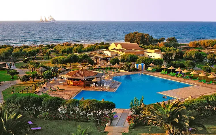 Hotelanlage mit großem Pool, grüner Gartenlandschaft und Blick auf das Meer bei Sonnenuntergang