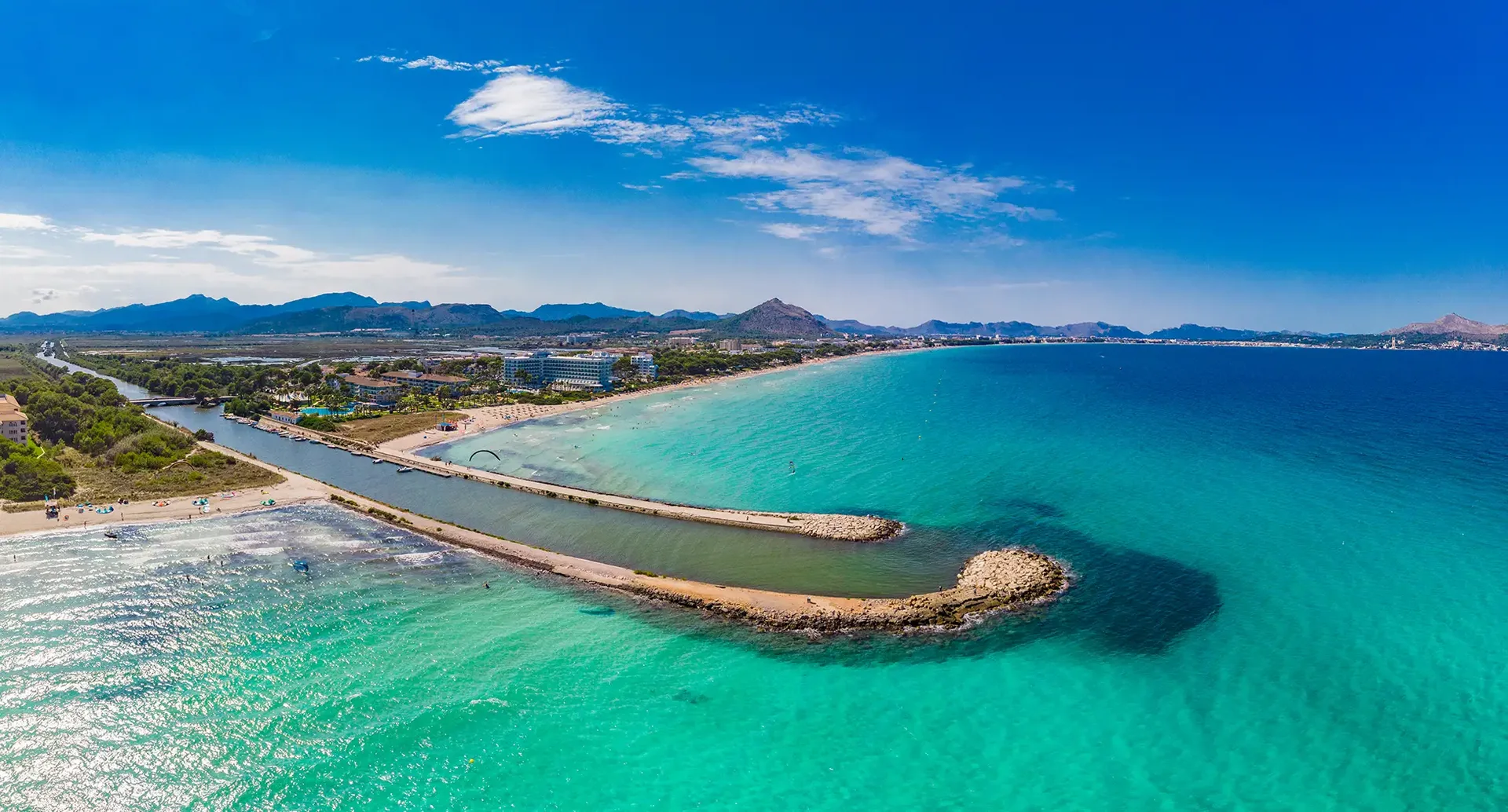 Weitwinkelaufnahme einer mallorquinischen Küste mit langer Sandbucht, klarem Wasser, geschwungener Mole und Berglandschaft im Hintergrund.
