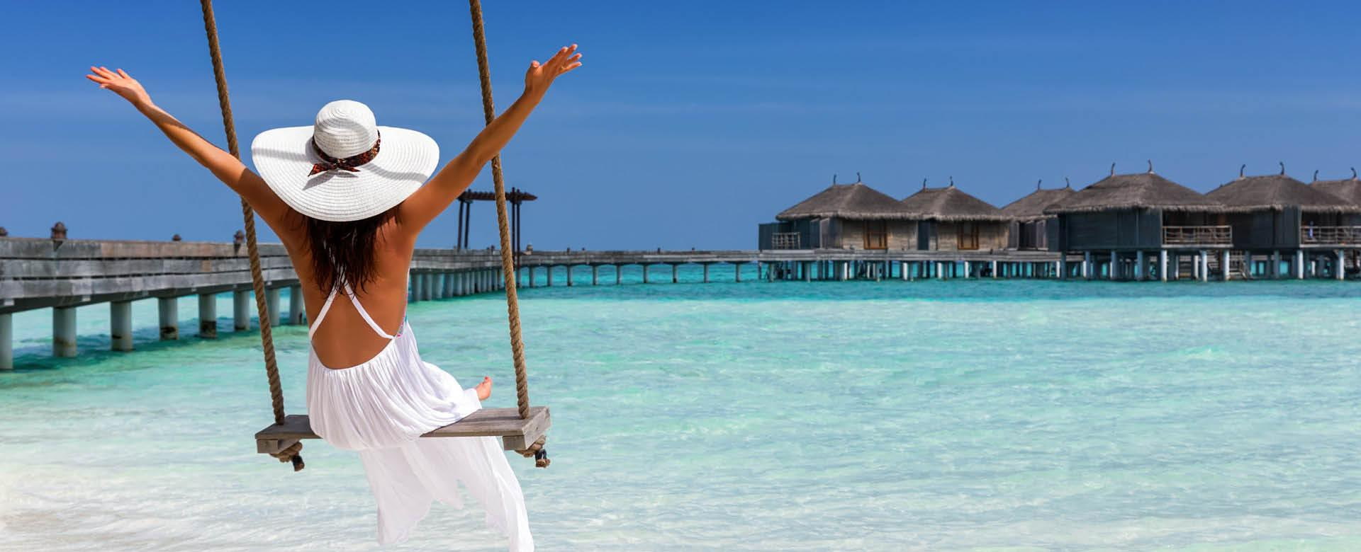 Frau im weißen Sommerkleid mit weißem Sonnenhut sitzt auf einer Schaukel am Strand mit ihren Armen in der Luft vor einem Wasserresort über dem türkisen Meer auf den Malediven