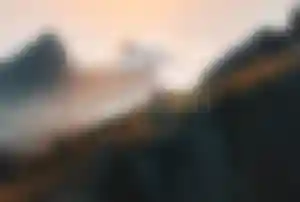 Person wandert auf einem schmalen Bergpfad entlang einer Felskante mit Blick auf eine weitläufige Berglandschaft bei Sonnenuntergang