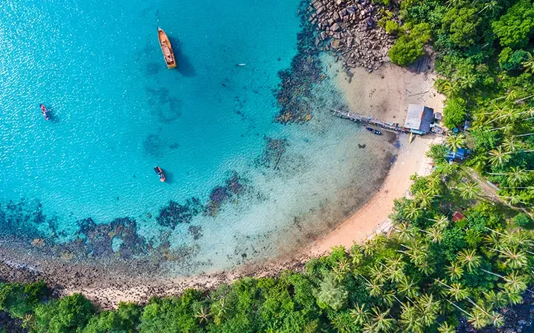 Luftaufnahme einer tropischen Bucht mit türkisblauem Wasser, kleinen Booten, Sandstrand und dicht bewachsenen Palmen am Ufer