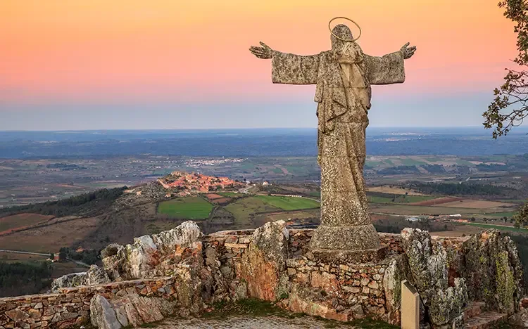 Steinerne Statue mit ausgebreiteten Armen auf einem Aussichtspunkt über einer weiten Landschaft mit Feldern, Hügeln und einem kleinen Ort im Hintergrund.