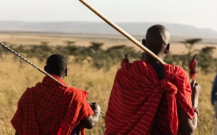 Zwei Menschen in traditionellen roten Gewändern gehen mit Speeren durch eine weite Savannenlandschaft mit vereinzelten Bäumen