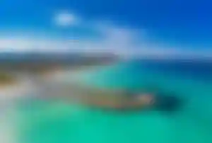 Panoramaaufnahme einer türkisblauen Bucht auf Mallorca mit geschwungener Landzunge, Sandstrand, Hotels im Hintergrund und Bergen am Horizont unter blauem Himmel.
