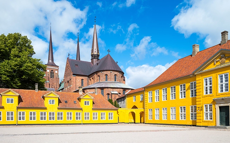 Blick auf den Dom von Roskilde mit roten Backsteinmauern und drei hohen Türmen, umgeben von gelben historischen Gebäuden.