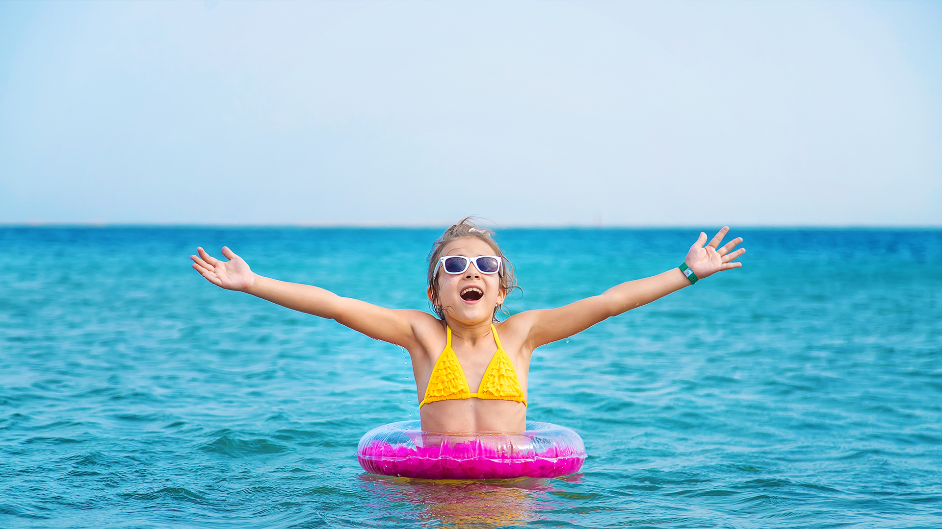 Mädchen in gelben Bikini hüpft vor Freude im Meer in ihrem rosa Schwimmreifen