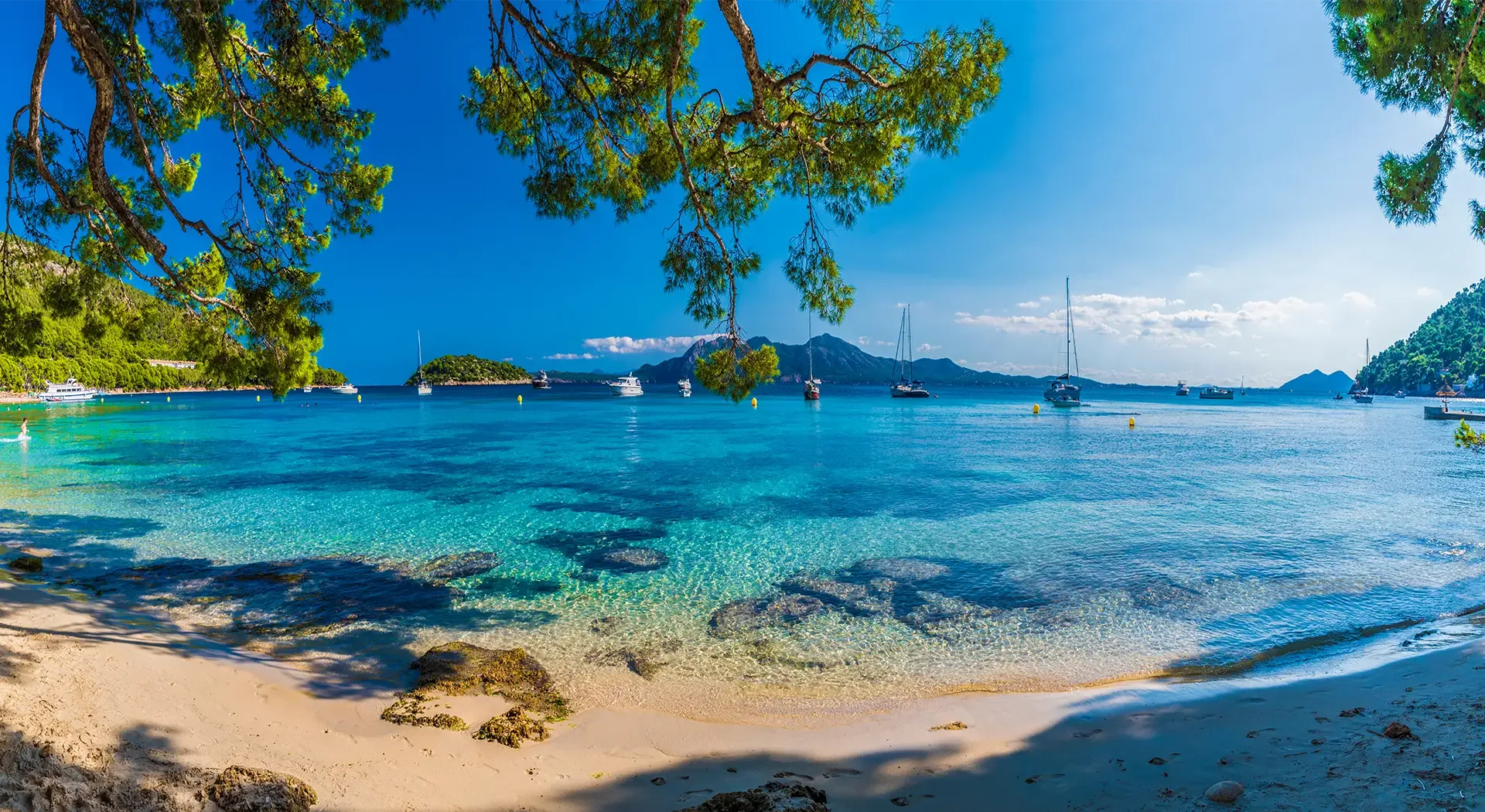 Sandstrand auf Mallorca mit kristallklarem Wasser, Pinien und vor Anker liegenden Segelbooten
