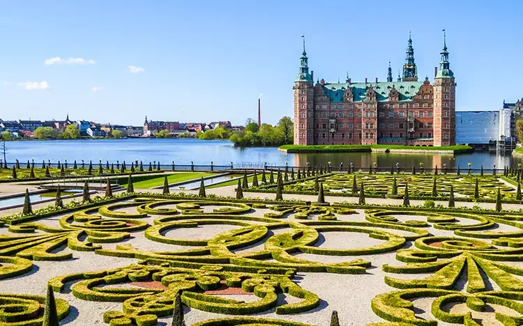  Frederiksborg Schloss mit grünen Türmen und Spiegelung im Wasser, davor kunstvoll angelegter Barockgarten.