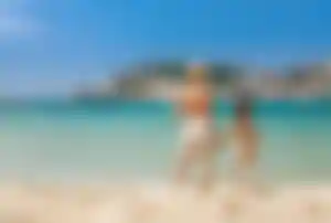 Mutter und Tochter stehen Hand in Hand am Strand auf Mallorca und Blicken aufs Meer