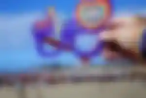 Hand mit Regenbogenarmband hält eine Pride-Brille vor dem Strand von Maspalomas auf Gran Canaria