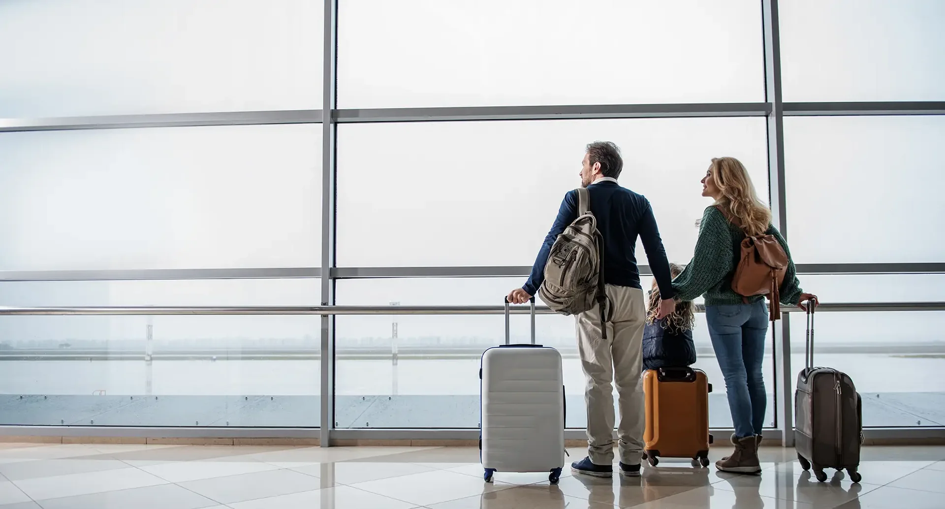 Familie steht mit Handgepäckskoffer vor großen Fenster am Flughafen