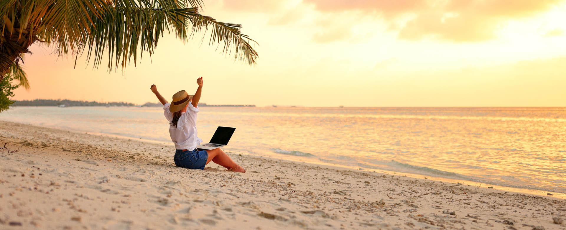 Frau sitzt mit Laptop an Strand und hat Arme vor Freude in der Luft.