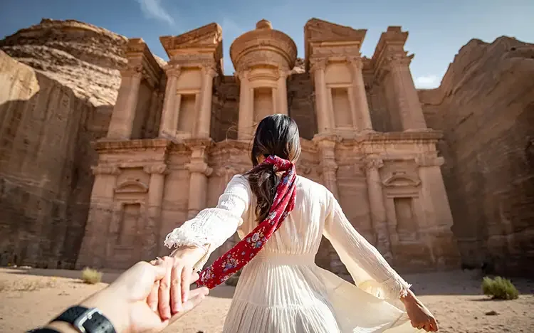 Person steht vor der monumentalen Fassade des Klosters in Petra und hält eine Hand nach hinten ausgestreckt.