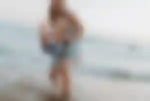 Person hält ein Kind im flachen Wasser am Strand, während Meerwasser aufspritzt und Wellen im Hintergrund zu sehen sind