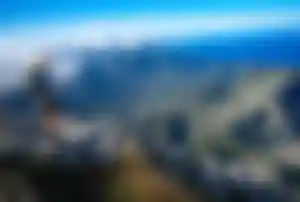 Person mit Rucksack steht auf einem felsigen Aussichtspunkt in der Serra de Tramuntana und blickt über eine bergige Landschaft bis zum Meer.