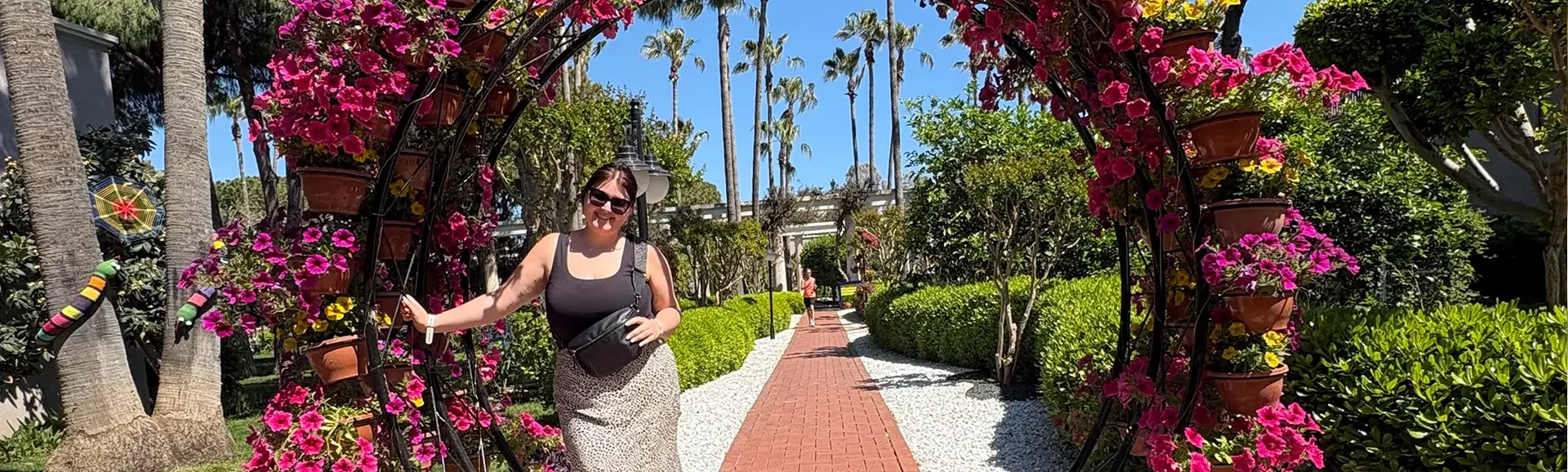 Person schreitet durch einen reich bepflanzten Blumenbogen in einer sonnigen Hotelgartenanlage in Antalya mit Palmen, Wegen und üppigem Grün.