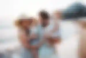 Vier Personen verbringen gemeinsam Zeit an einem Sandstrand am Meer, zwei Erwachsene halten jeweils ein kleines Kind auf dem Arm.