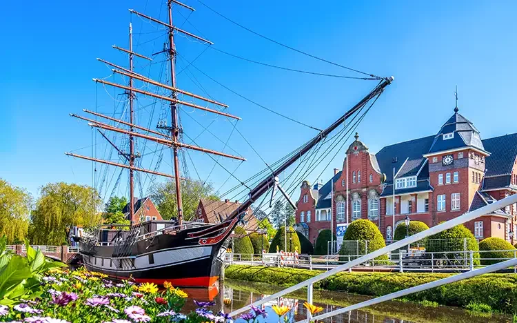 Historisches Segelschiff vor einem Backsteingebäude und Blumen am Kanal in Papenburg.