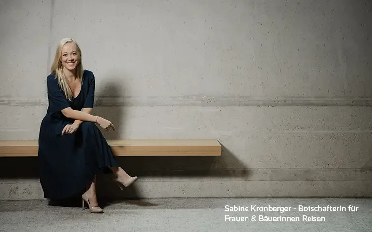 Person sitzt auf einer Holzbank vor einer hellen Betonwand; unten rechts ist der Text „Sabine Kronberger – Botschafterin für Frauen & Bäuerinnen Reisen“ sichtbar.