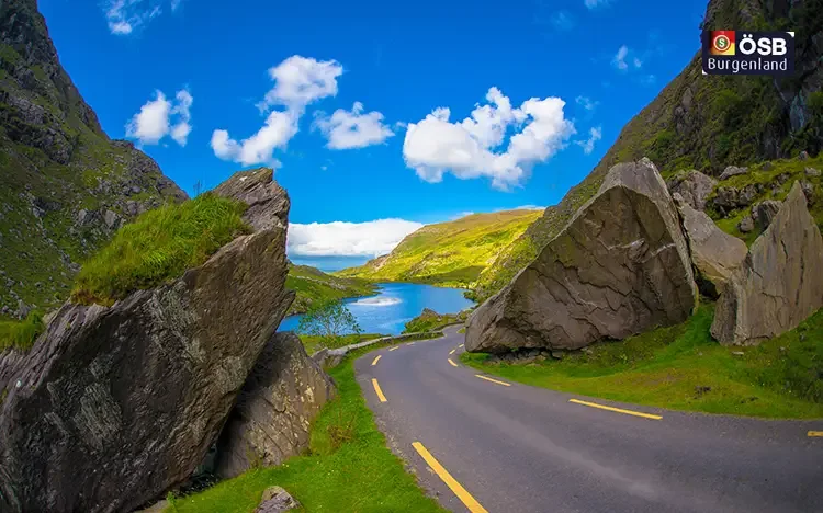 Malerische Straße am Ring of Kerry mit grünen Hügeln und Blick auf einen See unter blauem Himmel.