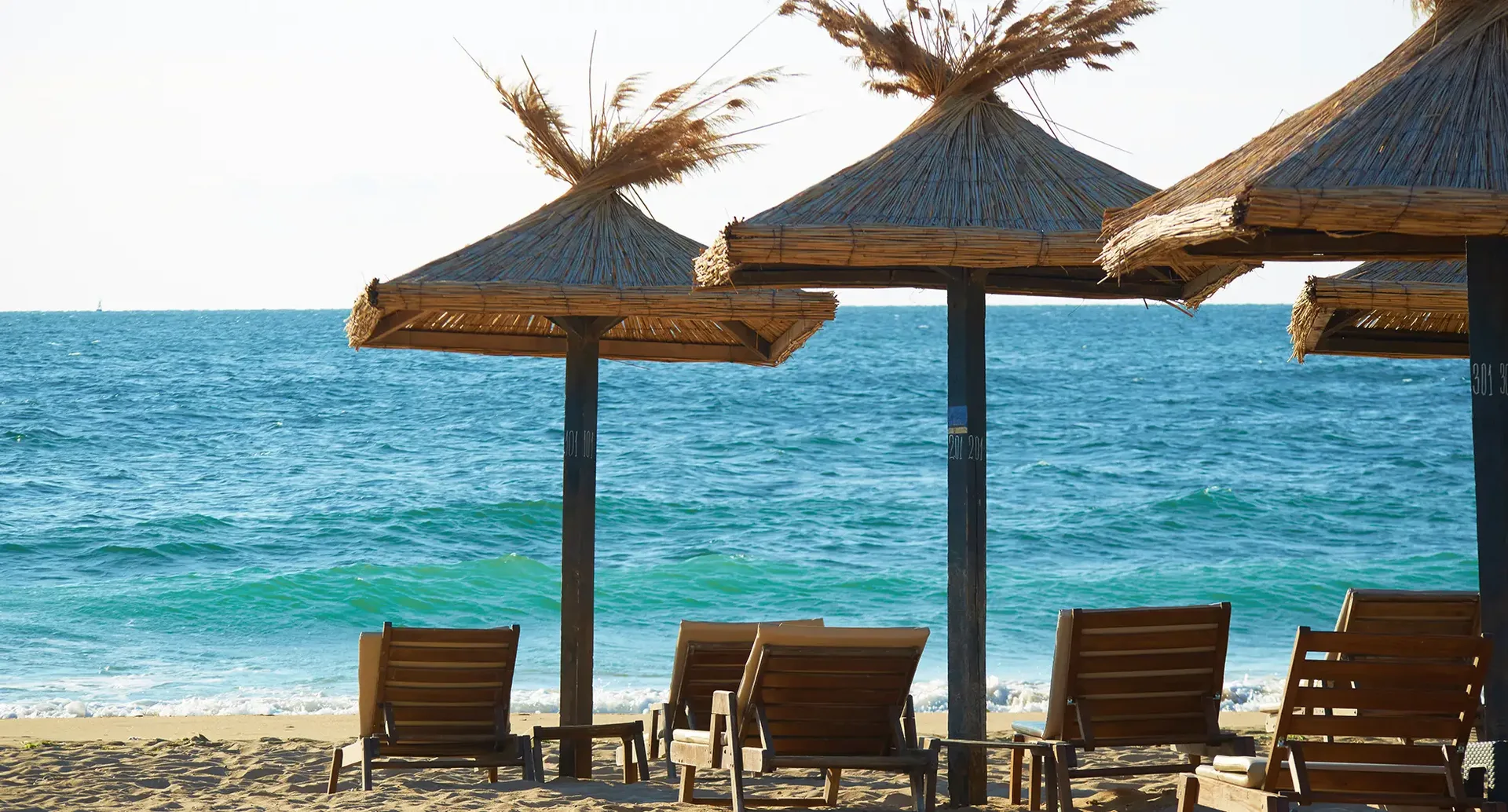 Strandliegen in Bulgarien – Entspannung am Schwarzen Meer Holzliegen und Strohdach-Sonnenschirme an einem Strand in Bulgarien mit ruhigem, blauem Meer im Hintergrund.