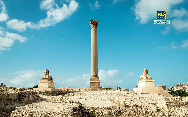 Pompeius-Säule in Alexandria, flankiert von zwei Sphinx-Statuen vor blauem Himmel